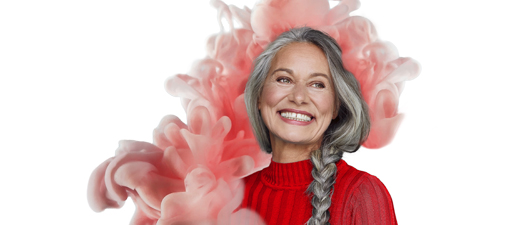 Bildbeschreibung: Eine ältere lachende Frau mit langen grauen Haaren, die zu einem Zopf geflochten sind. Sie trägt einen roten Pulli und steht vor einer künstlichen rosafarbenen Wolke, die wie Watte aussieht.