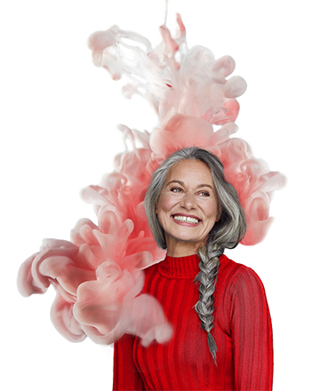 Bildbeschreibung: Eine ältere lachende Frau mit langen grauen Haaren, die zu einem Zopf geflochten sind. Sie trägt einen roten Pulli und steht vor einer künstlichen rosafarbenen Wolke, die wie Watte aussieht.