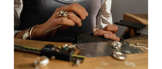 Bildbeschreibung: Auf dem Foto ist eine junge Goldschmiedin zu sehen, die mit Goldschmiedewerkzeugen einen goldenen Ring mit einem eingefassten Schmuckstein bearbeitet. Die Frau hat blonde zu einem Zopf gebundene Haare, trägt eine grau-weiß gestreifte Bluse und wirkt sehr konzentriert bei der Arbeit. Am rechten Ringfinger trägt sie einen sehr auffälligen Ring. Außerdem trägt sie eine goldenen Kette und einen goldenen Armreif sowie eine Armbanduhr am rechten Arm.