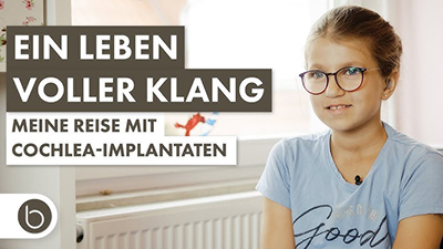Bildbeschreibung: Das Bild zeigt einen Ausschnitt aus dem Film Meine Reise mit Cochlea-Implantaten. Darauf abgebildet ist die Protagonistin, die elfjährige Lena Pohl. Ihr Cochlea-Implantat ist durch die Haare verdeckt. Lena trägt eine Brille und ein hellblaues T-Shit sowie Ohrstecker, sie lächelt. Ihre Haare sind zu einem Zopf zusammengebunden.