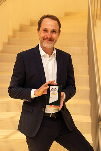 Bildbeschreibung: Auf dem Bild ist Gert Ettlmayr abgebildet. Er ist Vertriebschef und Senior Director Sales & Operations bei Geers. In beiden Händen hält er den Deutschen Service-Preis 2026. Ettlmayr ist mittleren Alters, er hat braune kurze Haare, eine hohe Stirn und einen graumelierten Bart und Schnurrbart. Er lächelt direkt in die Kamera. Er trägt einen schwarzen schicken Anzug und ein weißes Hemd.