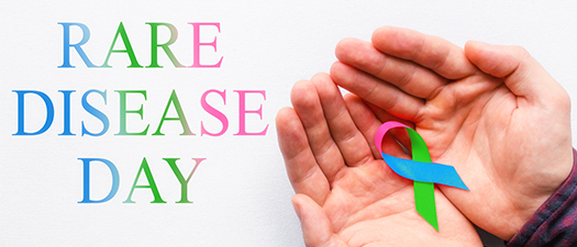 Bildbeschreibung: Auf der linken Seite des Fotos steht in buten Großbuchstaben das Wort Rare Disease Day. Auf der rechten Seite sind zwei nebeneinander liegende Hände abgebildet, in deren Handflächen eine bunte Schleife liegt.