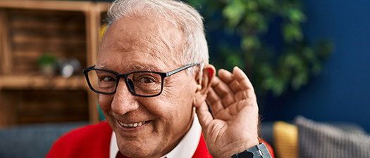 Bildbeschreibung: Auf dem Bild ist ein älterer Herr zu sehen. Er lächelt und hält eine Hand hinters Ohr.