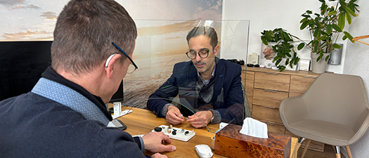 Bildbeschreibung: Auf dem Foto ist ein Hörakustiker in seinem Fachgeschäft zu sehen, der einem Kunden mehrere Hörsysteme vorstellt.