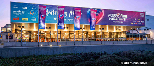 Auf dem Foto ist die Wiener Stadthalle von außen zu sehen. Mit Fahnen und einem großen Banner wird auf den dort stattfindenden 70. Eurovision Song Contest hingewiesen.