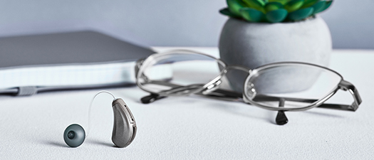 Auf dem Foto ist ein Hörsystem zu sehen. Dahinter befindet sich, zum Größenvergleich, eine Brille.