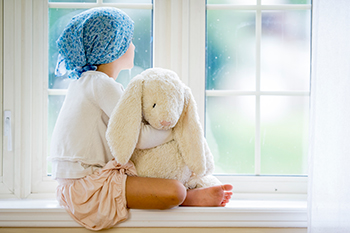 Das Bild zeigt ein krebskrankes Kind, das einen Teddy im Arm hat und aus dem Fensterschaut